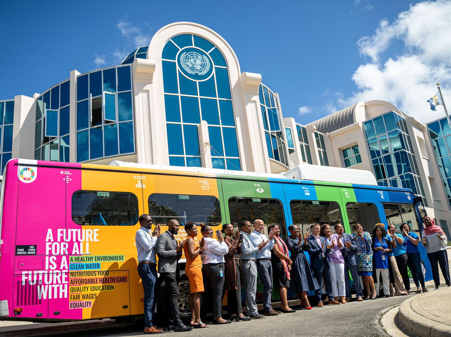 group of 15 persons stand together applauding for a photo against a bus wrapped brightly in the SDG colours, with the UN Seal on a building beyond the bus