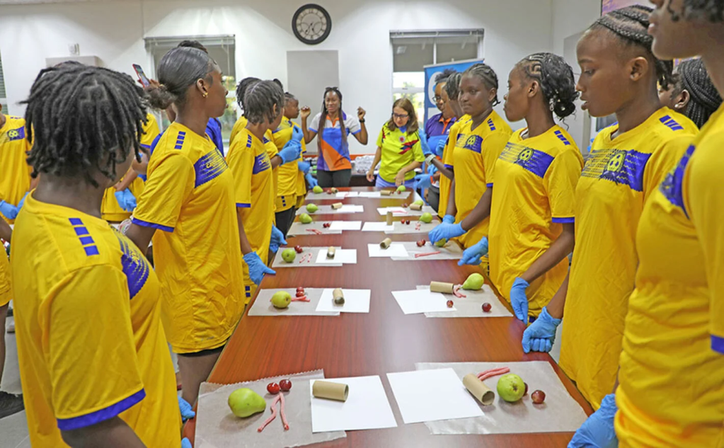 a group of girls in football uniforms lining the sides of a long table looking at the facilitator at the head table