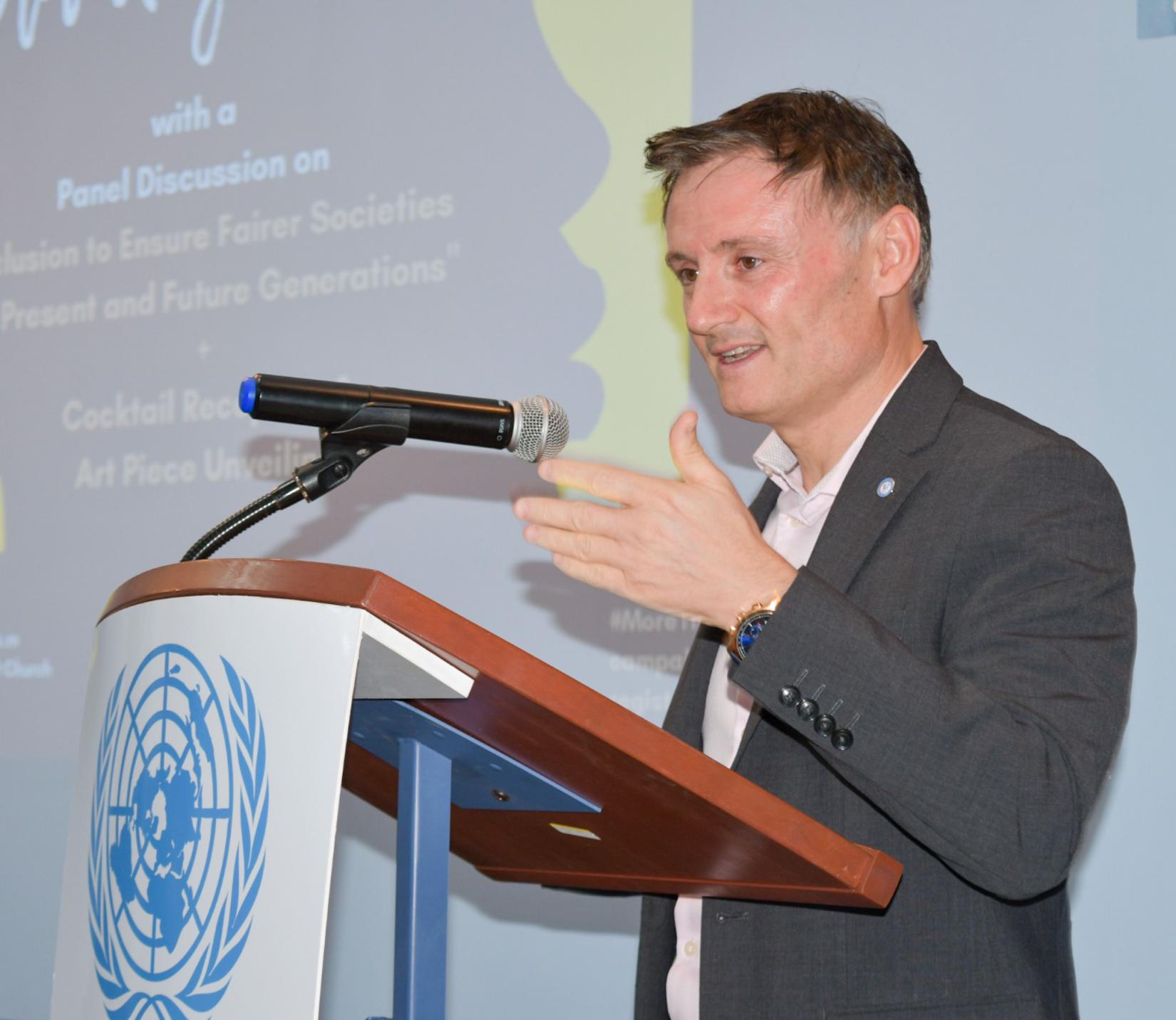 UN Resident Coordinator, Didier Trebucq stands at the UN podium on stage, delivering his remarks to the audience.