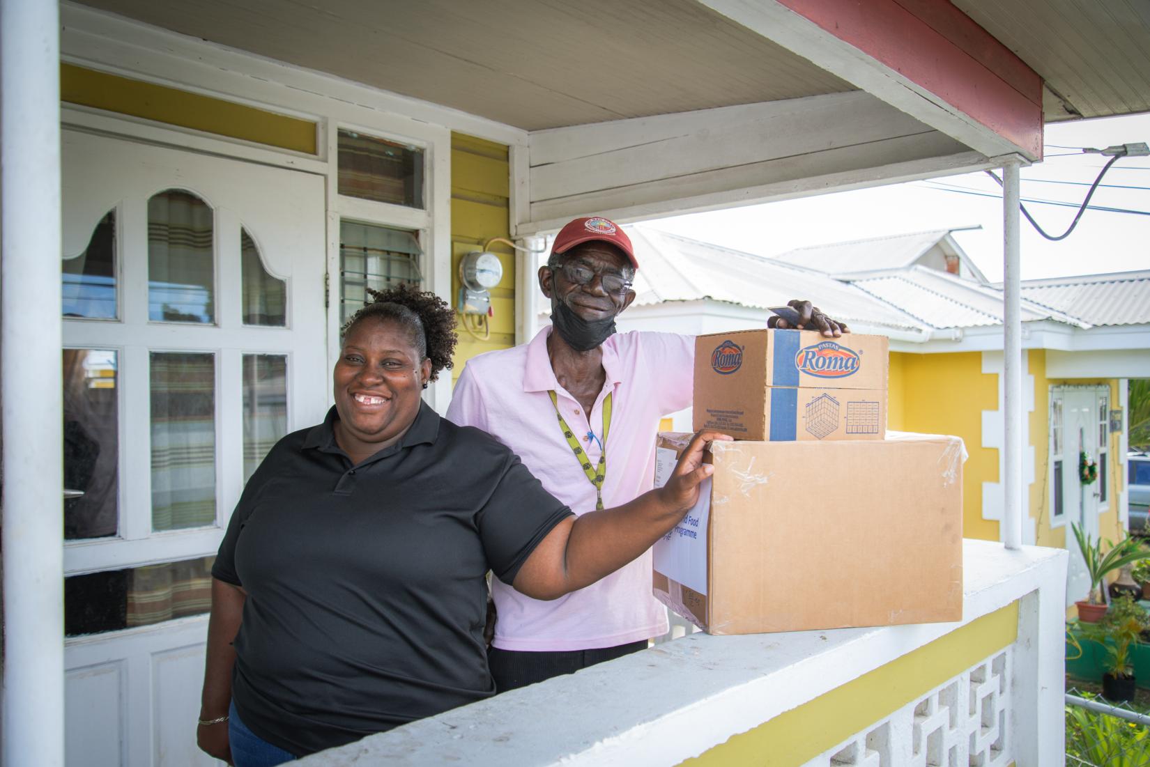 A smiling woman in a black polo shirt and a tall elderley man in a pink shirt, cap and mask beneath the chin stand with two boxes from the hamper drive. 