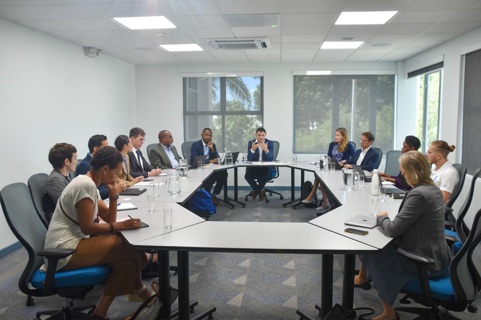 Image of persons sitting in a carpeted room in office chairs, around a hexagonal table focusing on the man sitting at the head of the table.