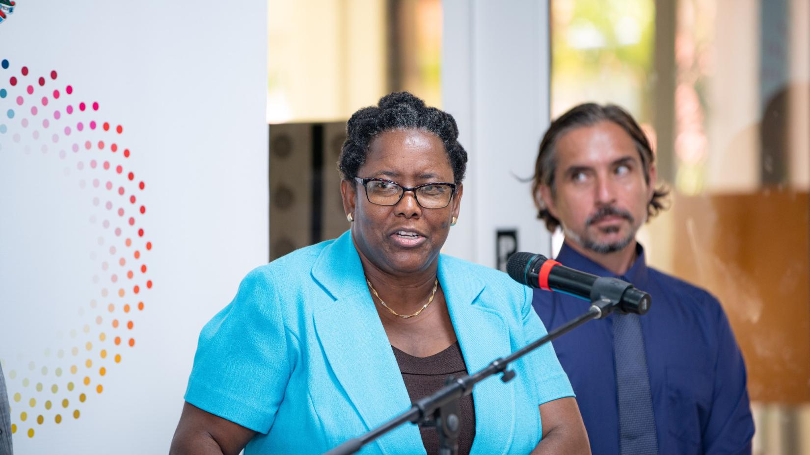 Woman at podium speaking into microphone to an audience with a banner behind her