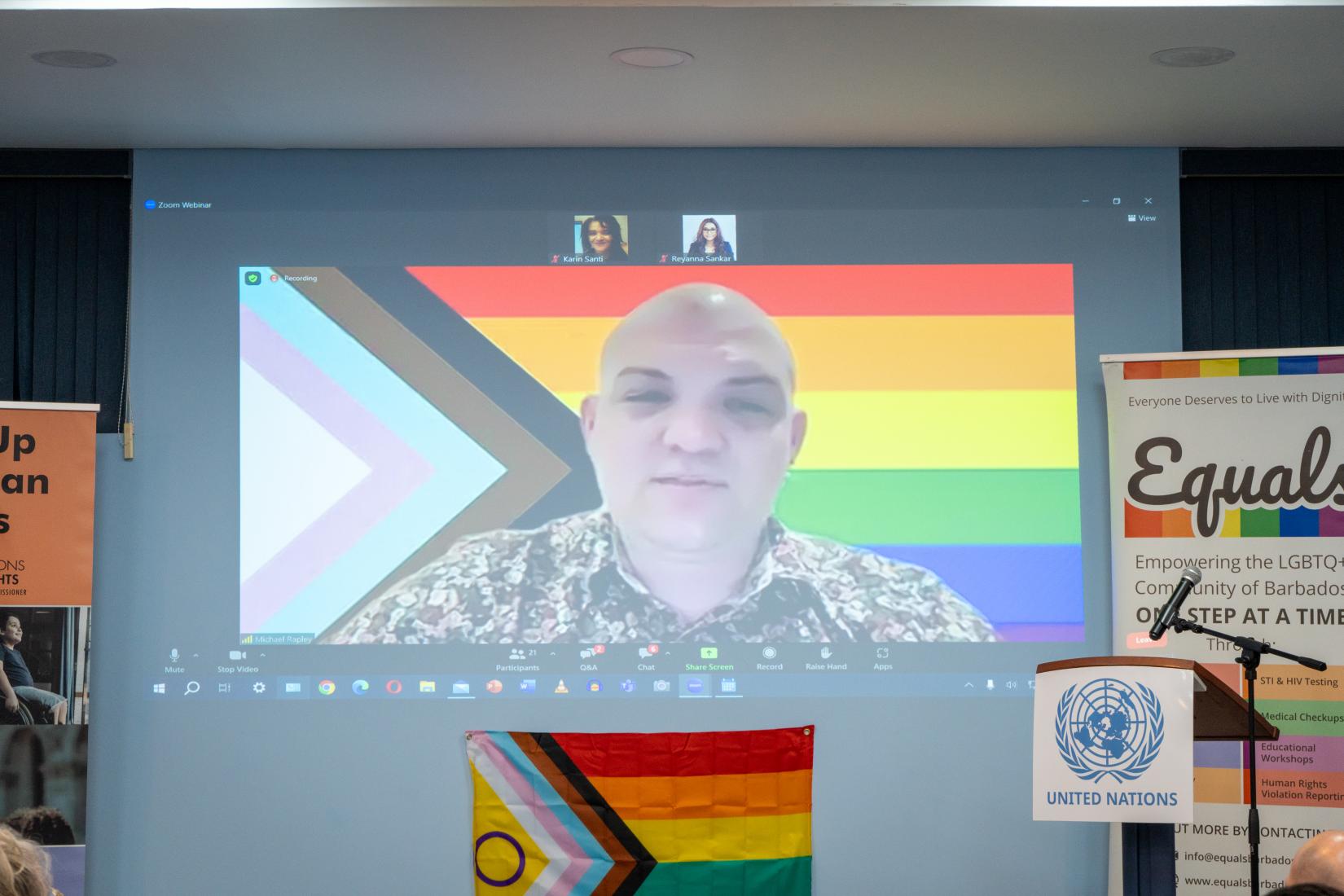 Man on a Zoom screen with a background of the LGBTI+ flag addressing an audience