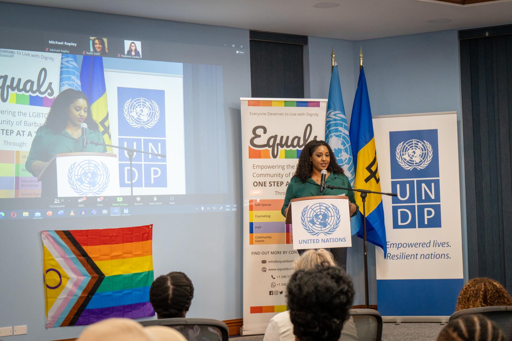 Lady stands on a stage at a podium before an audience with a projected Zoom screen to the left of her