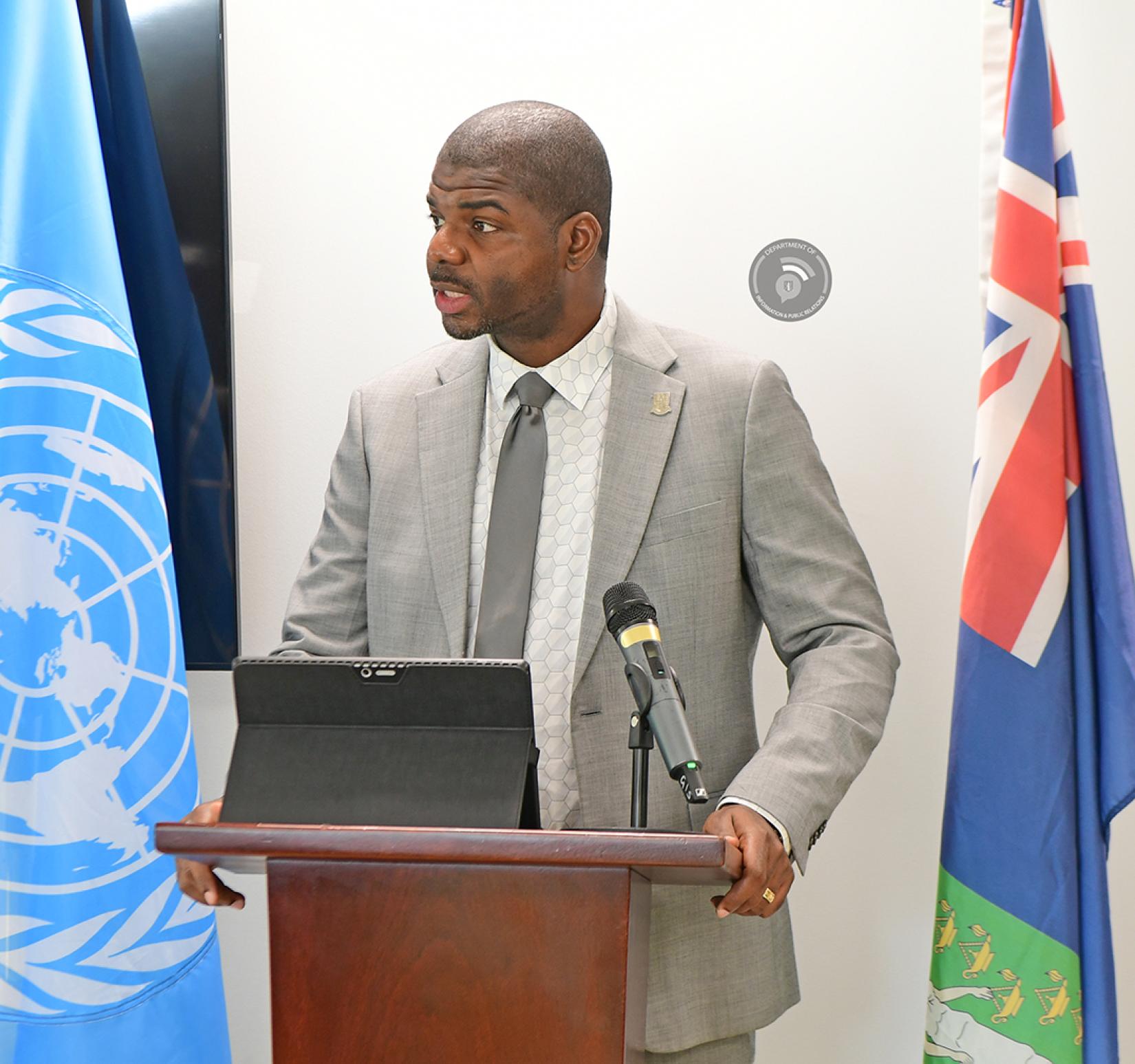 Man standing in grey suit at a podium between flags looks at audience with tablet in stand mode