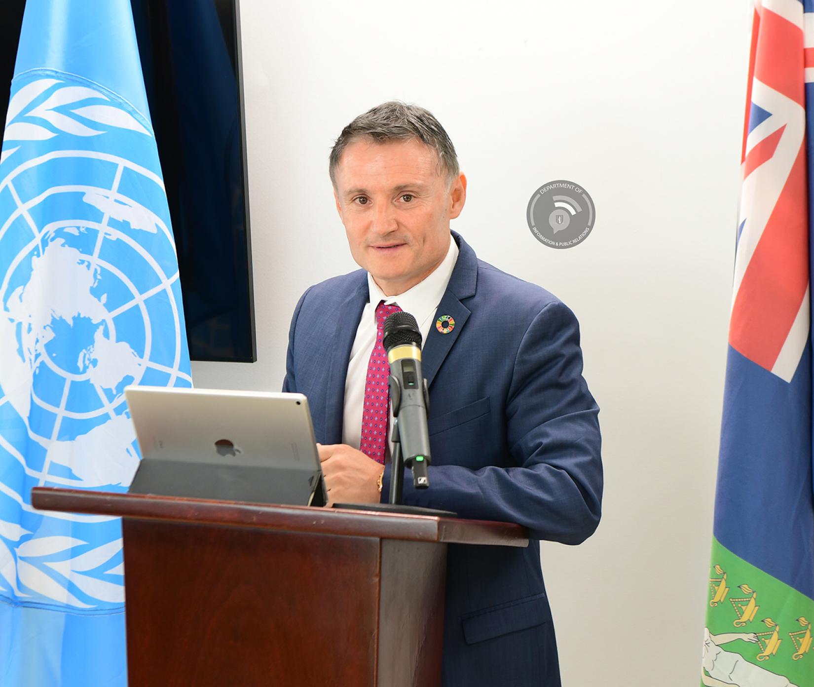 Man standing in navy blue suit at a podium between flags looks at audience with tablet in stand mode