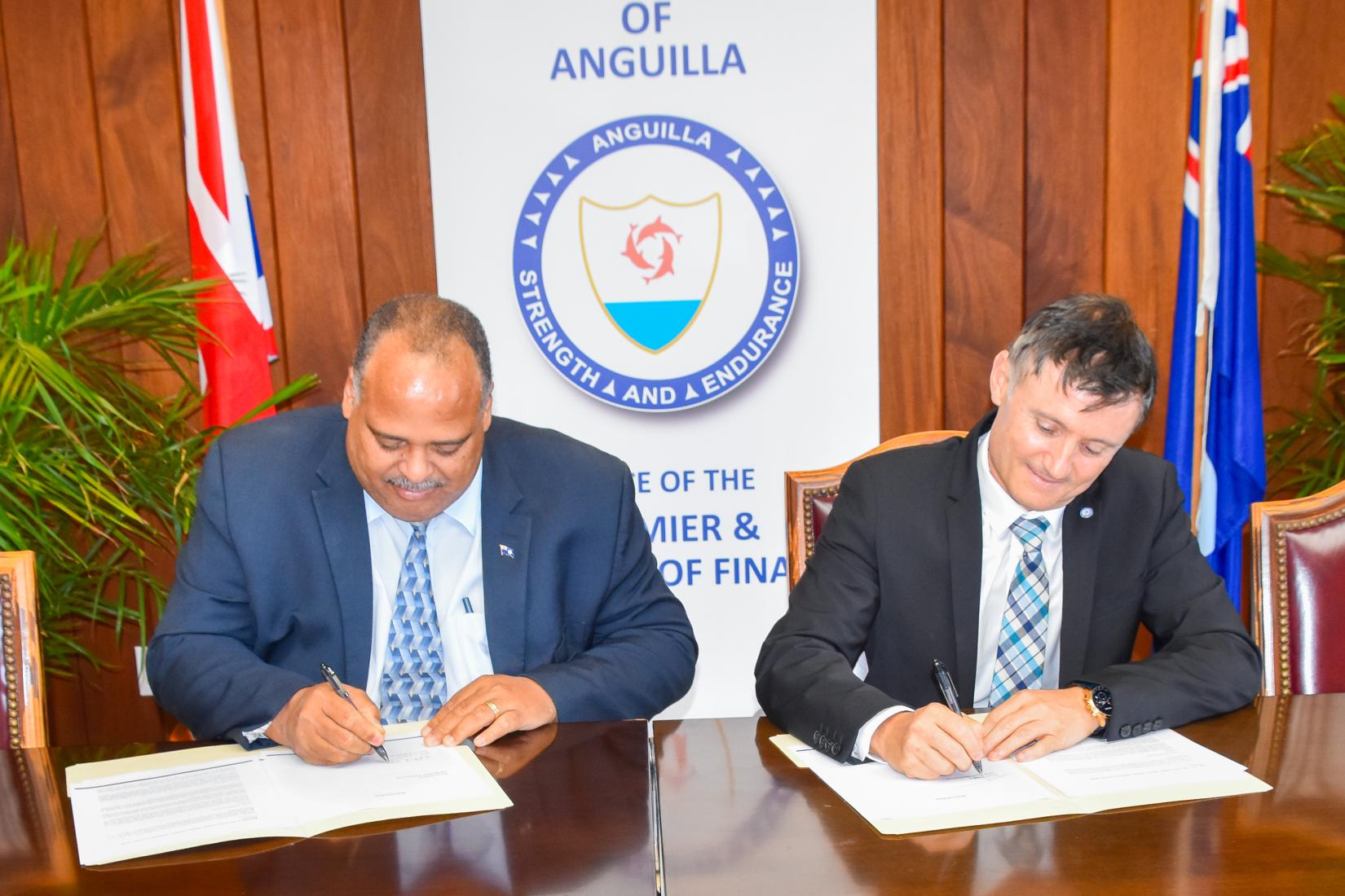 two men in suits sit beside each other at a mahogany table and sign copies of documents