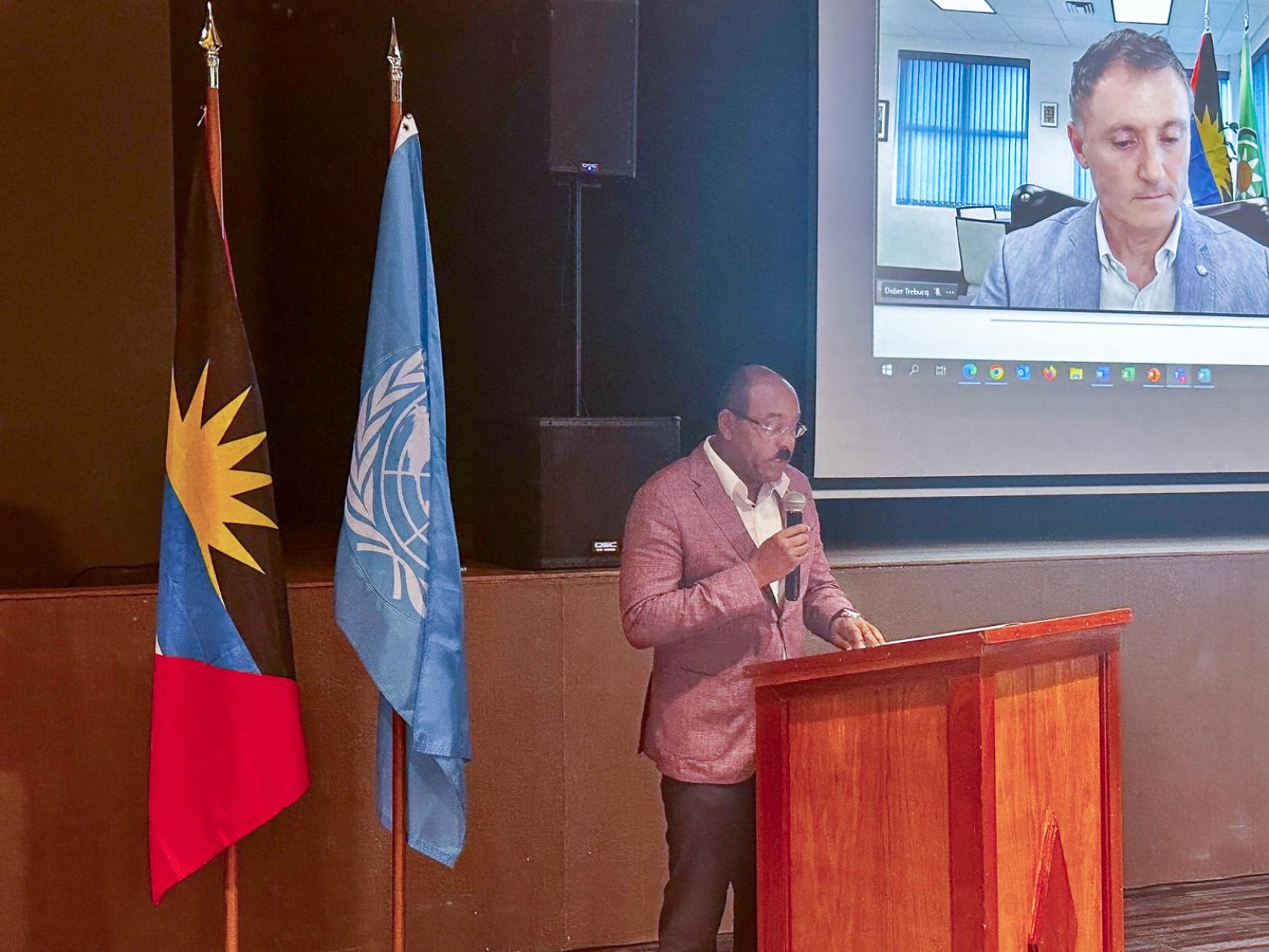 Man standing in blazer at podium with man on screen behind him, and two flags behind him.