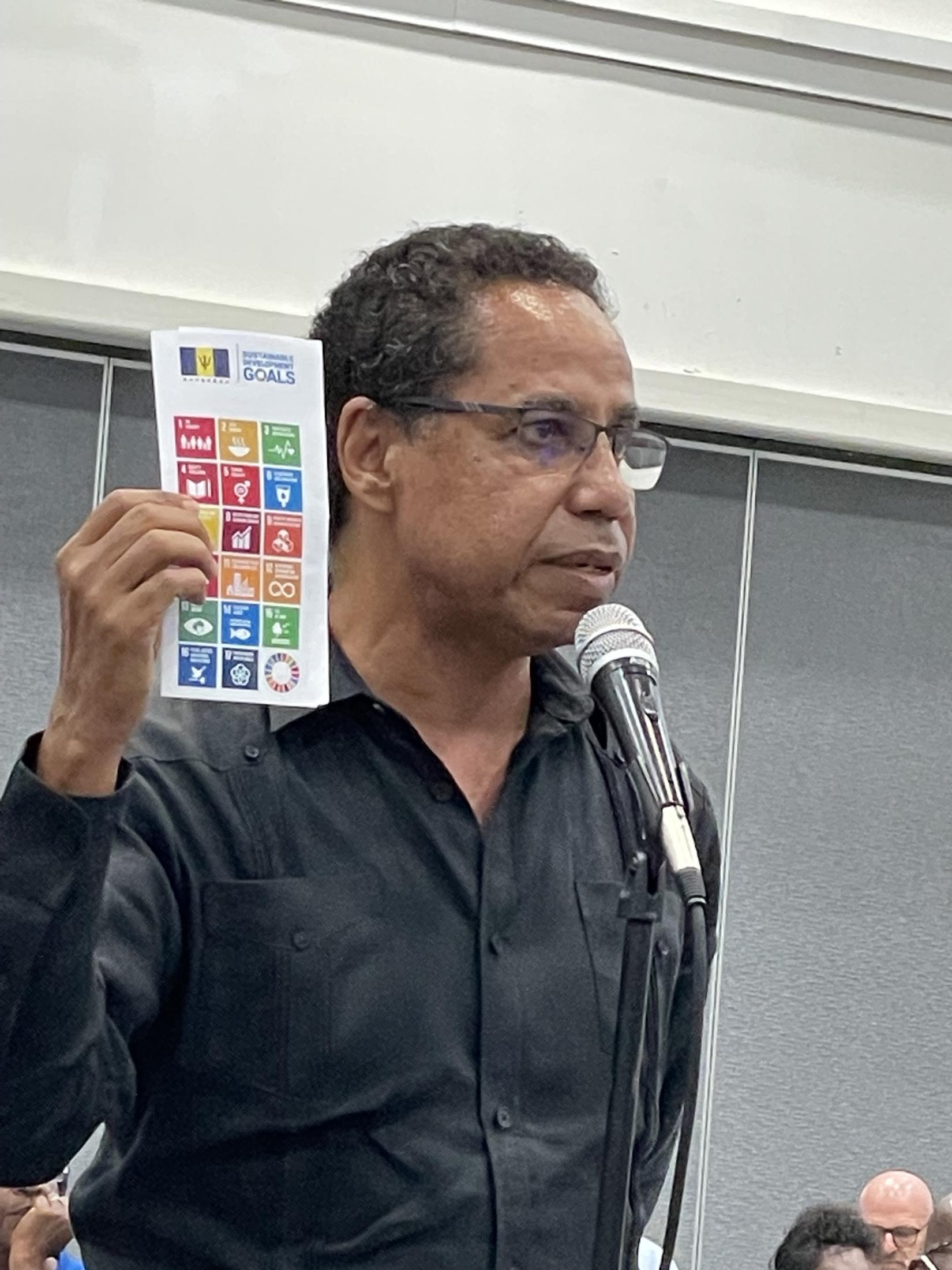 Man standing in audience at a microphone on a stand holding up a sheet of the Sustainable Development Goals