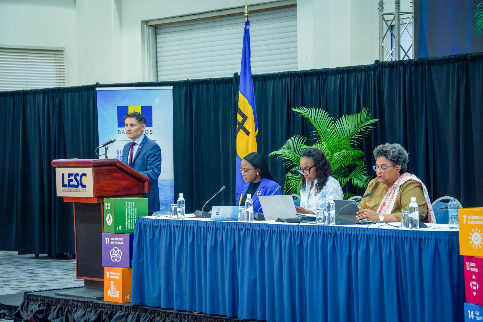 Man standing at podium facing the audience, with a panel to his right sitting in a large conference room