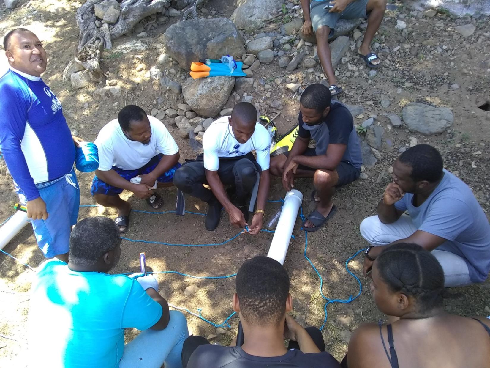 A group of people crouch in a dry riverbed while an instructor demonstrates a piece of equipment