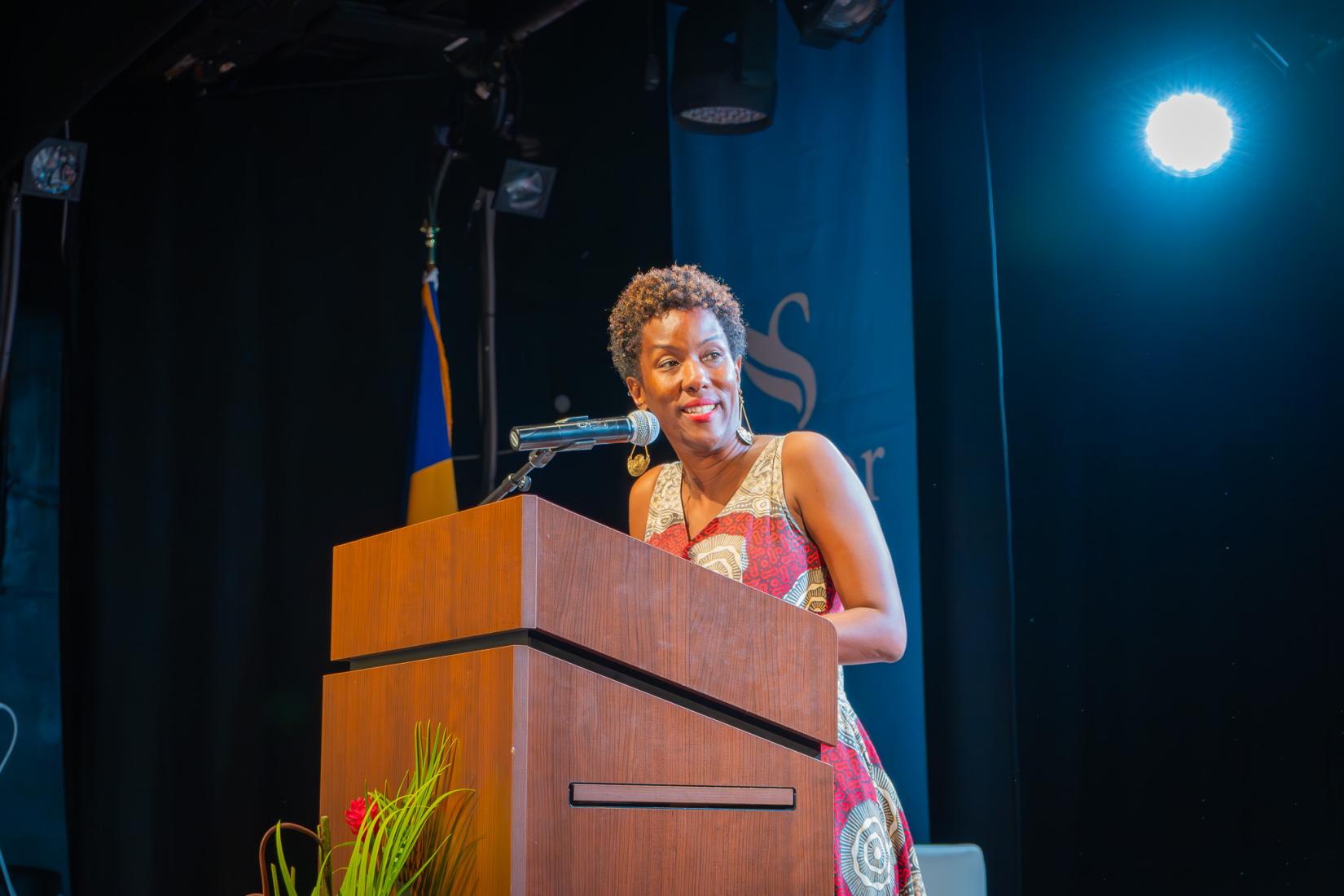 Woman at podium speaking into microphone to an audience with a banner behind her