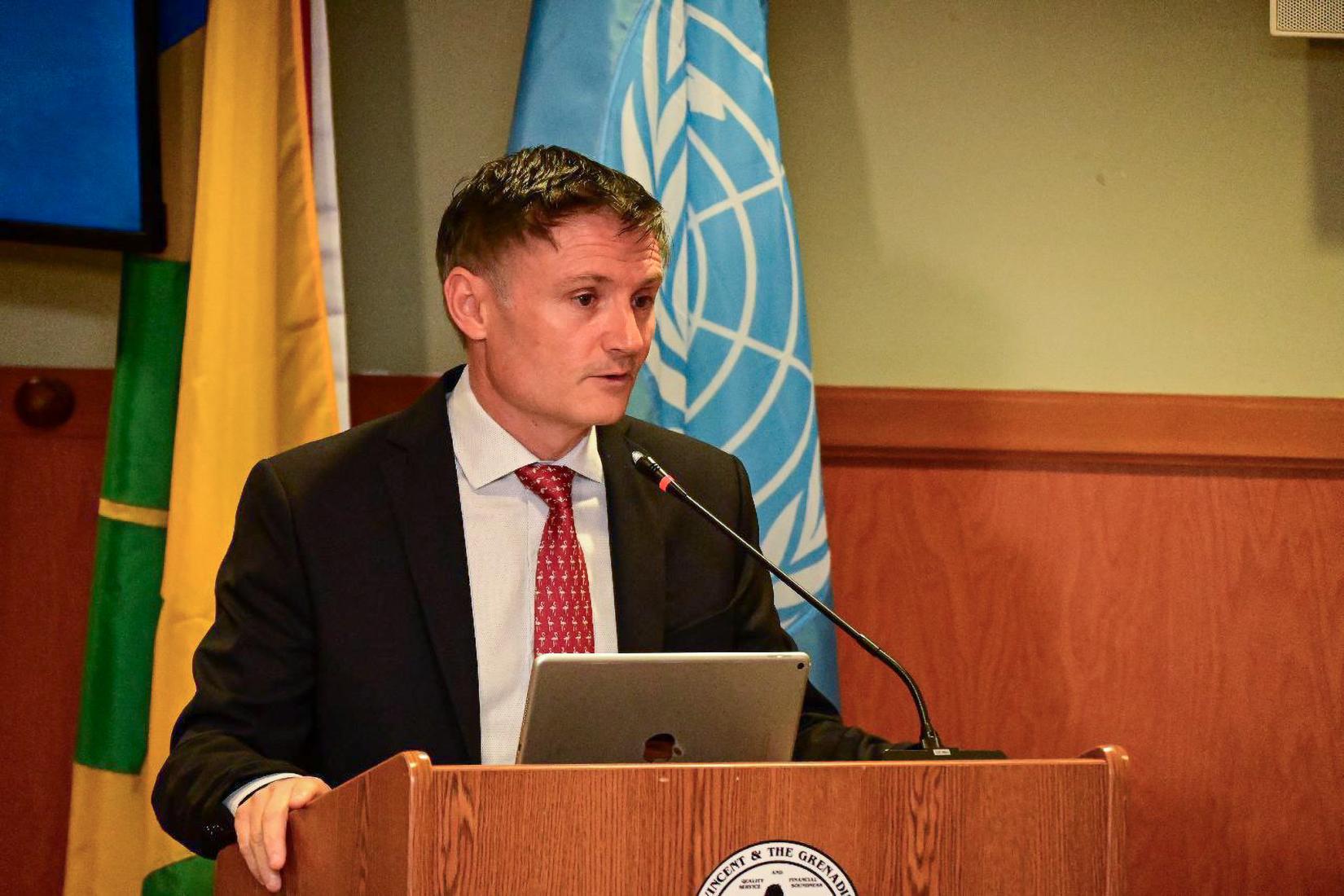 Man at podium speaks to an audience with flags in the background