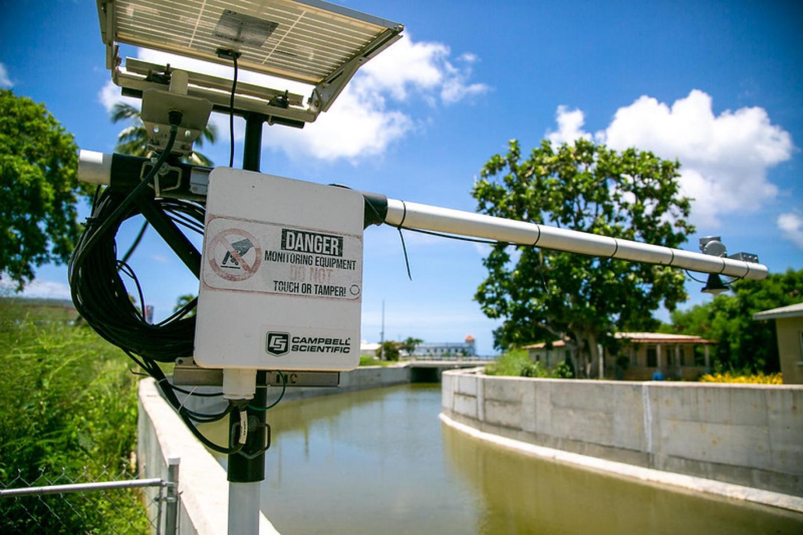 a piece of monitoring equipment in focus, along a waterway in a northern community in Barbados