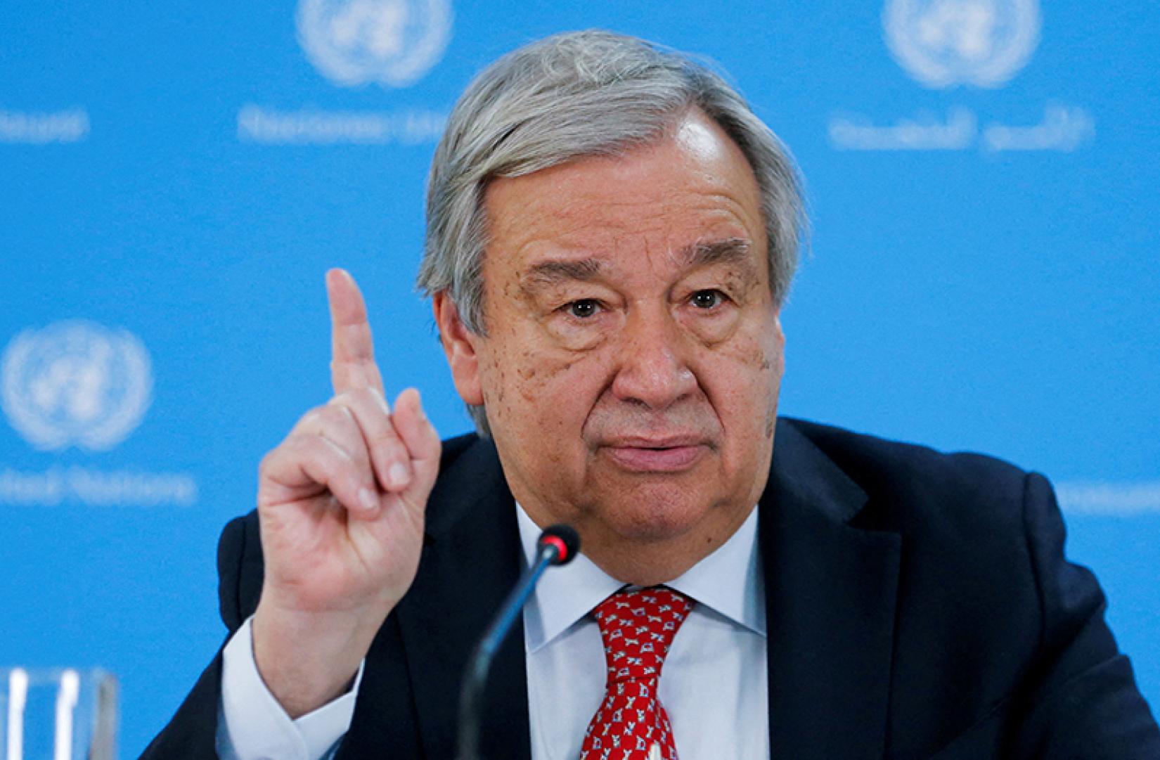 Photo of UN Secretary General mid speech, raising a finger with a UN Step and repeat banner blurred in the background 