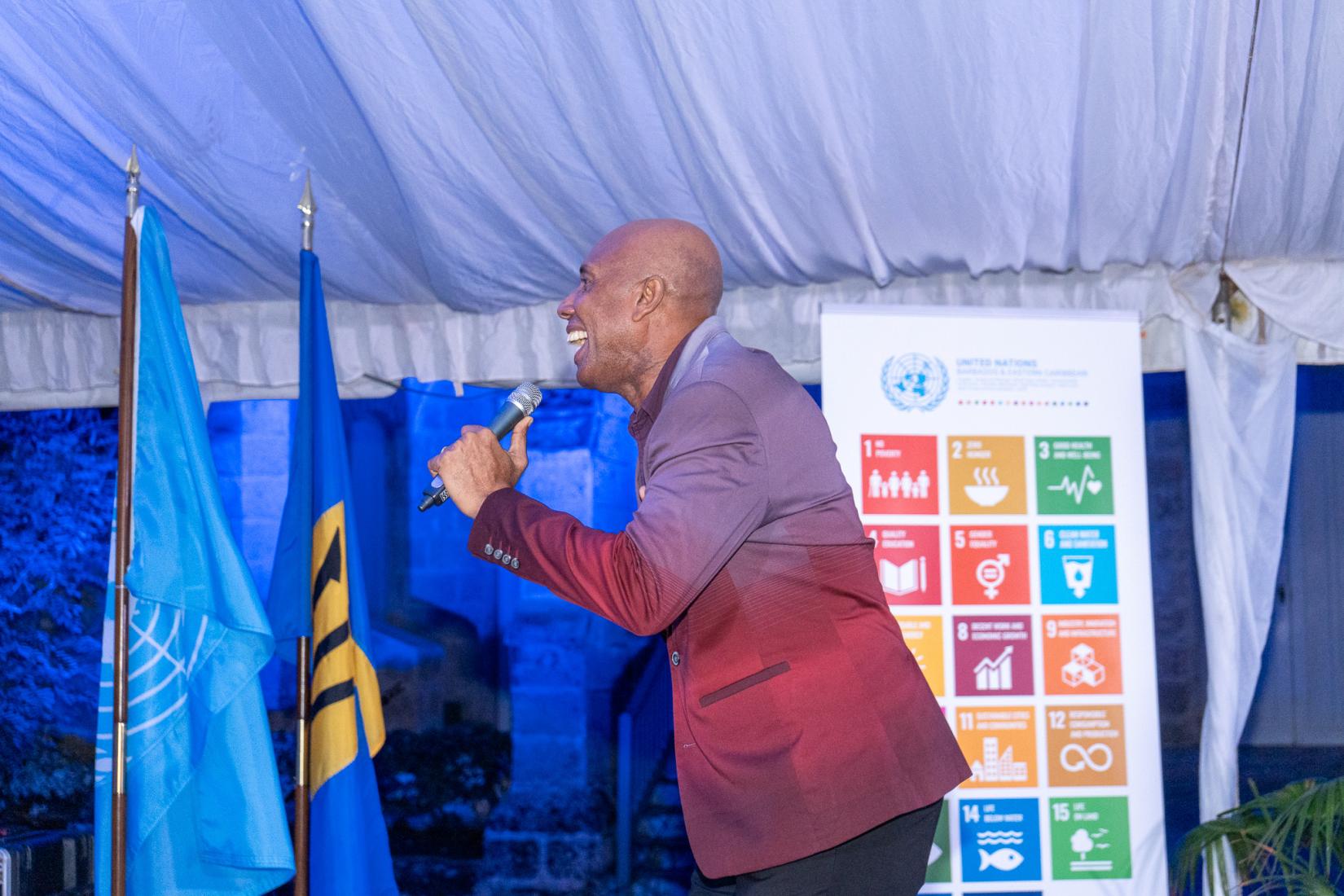 Man stands on stage in blazer with microphone behind him with the UN Flag, Barbados Flag, and standing banner of the 17 SDGs behind him