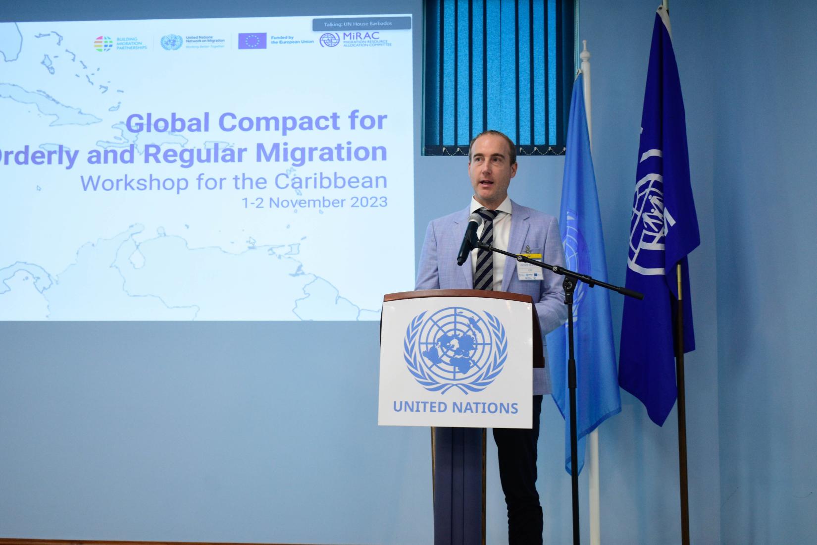 Man standing in blazer at podium with man on screen behind him, and two flags behind him.
