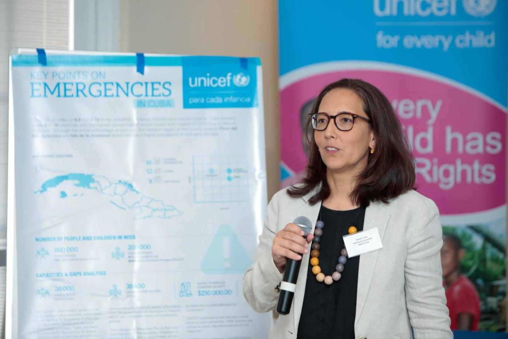 woman with microphone standing speaking to an audience with a chart on emergencies behind her, and a UNICEF banner in the background from behind a table