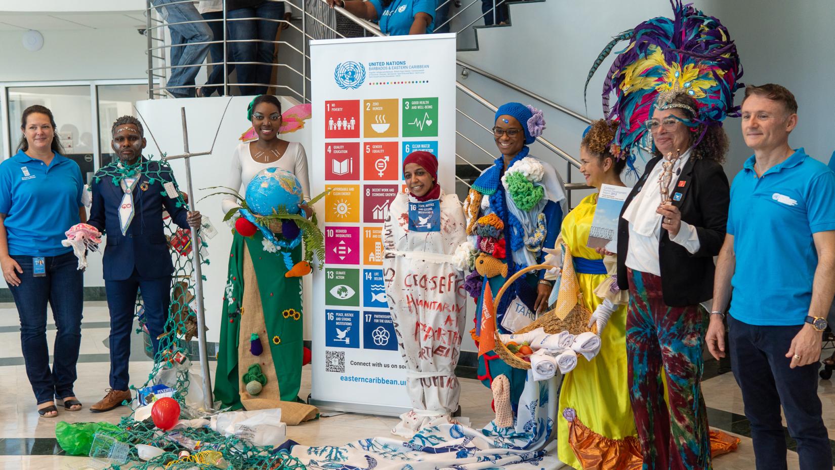group of people stand in colourful SDG themed costumes in front of a standing banner of the Sustainable Development Goals