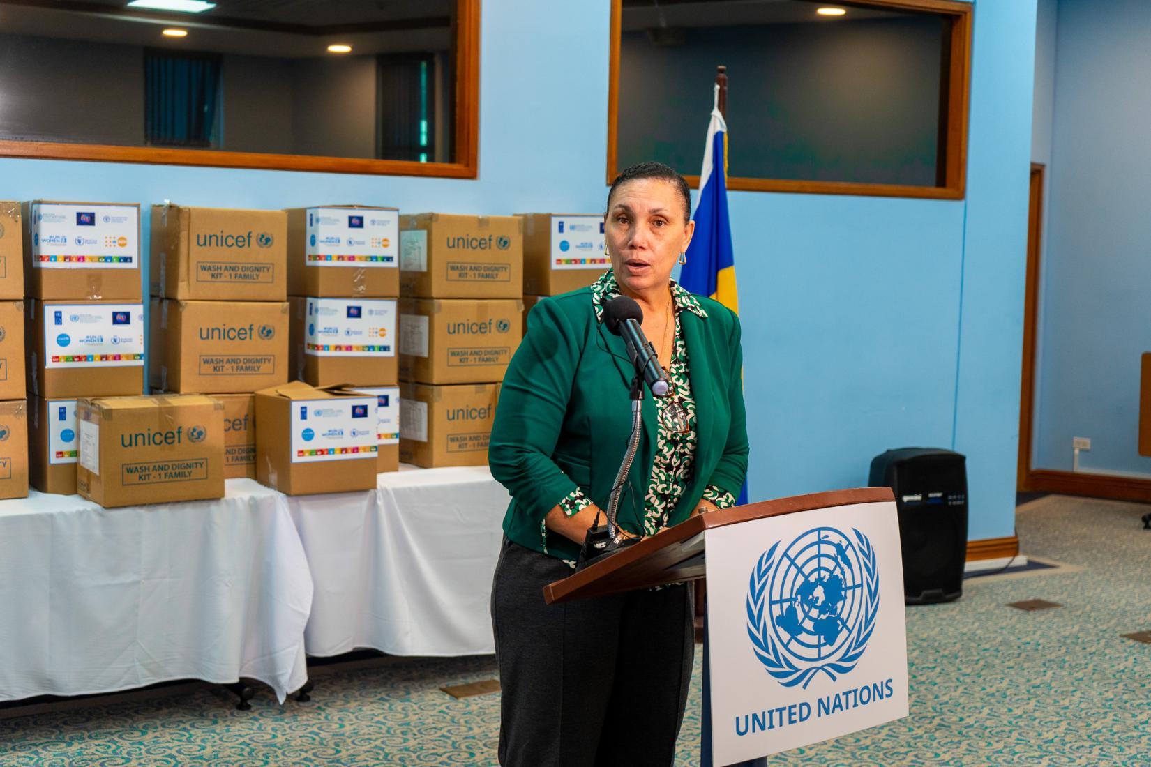 Woman at podium speaking into microphone to an audience with boxes behind her