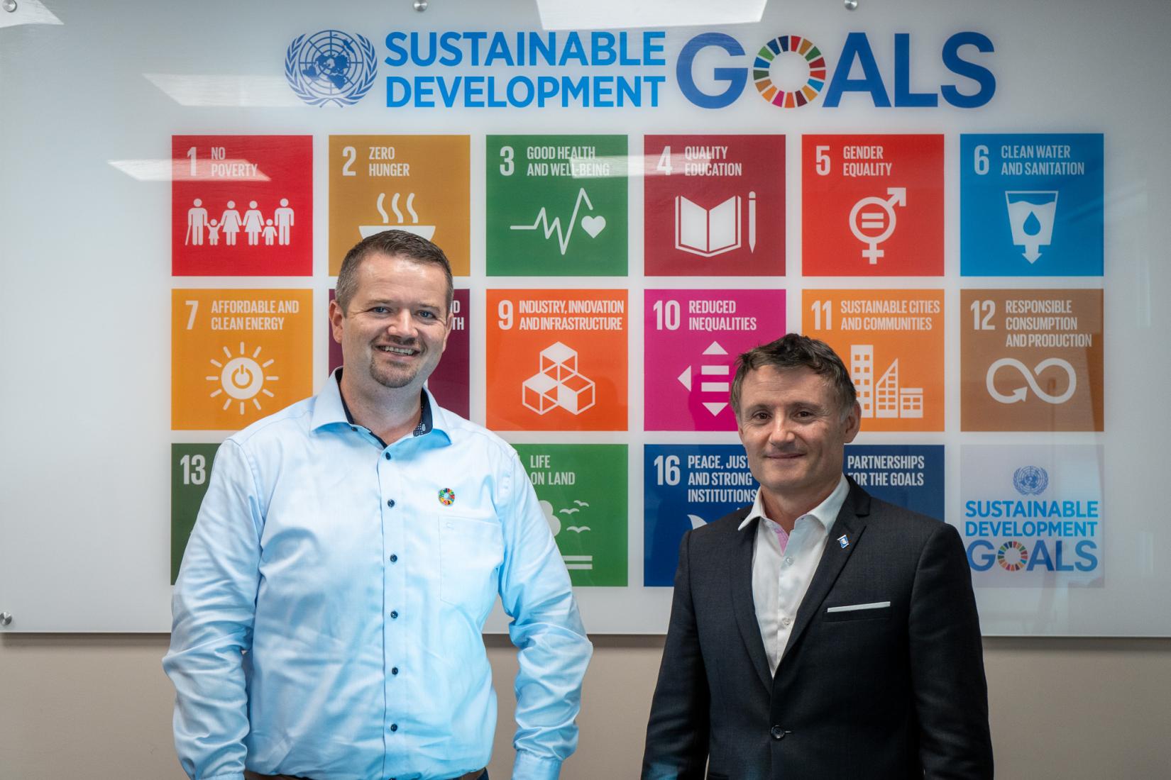 two men stand in front of a Sustainable Development Goals board in business wear