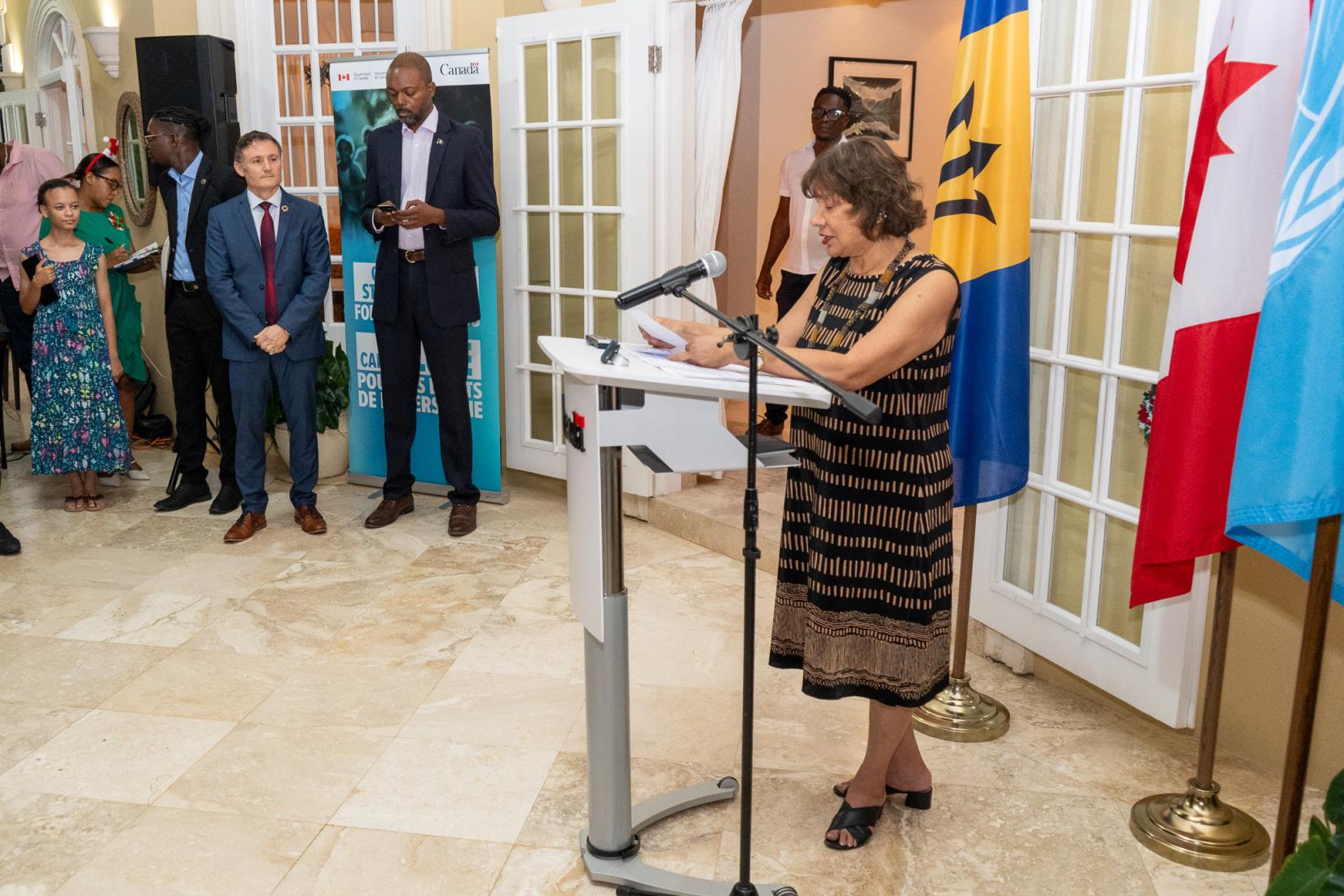 Woman at podium speaking into microphone to an audience with flags behind her. 