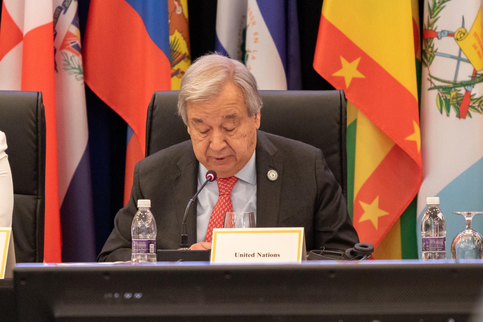 Man at table in suit speaks to an audience with flags in the background