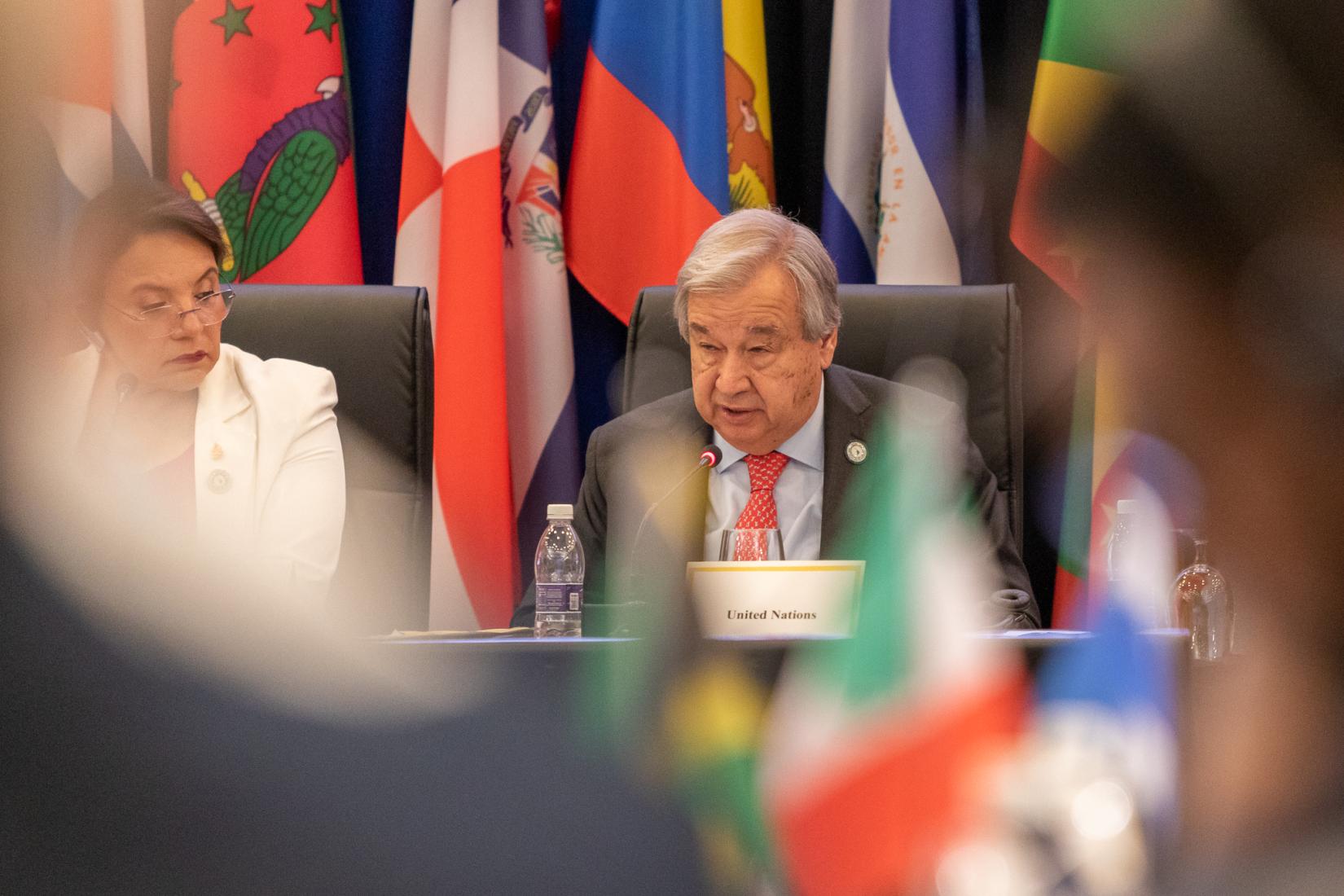 Man at table in suit speaks to an audience with flags in the background