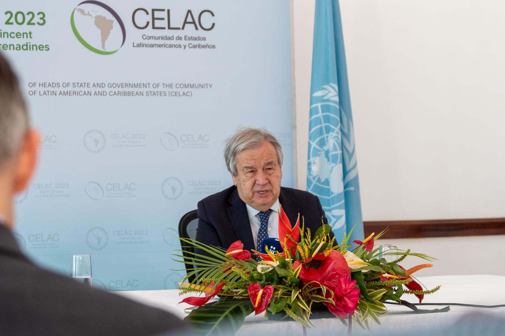 Man at table speaks to an audience with UN Flag in the background