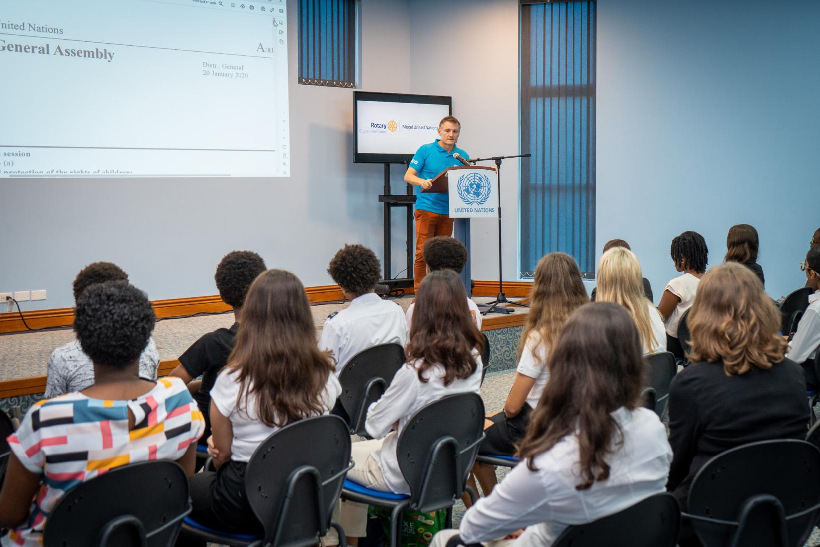 Man at podium on stage speaks to an audience of young children 