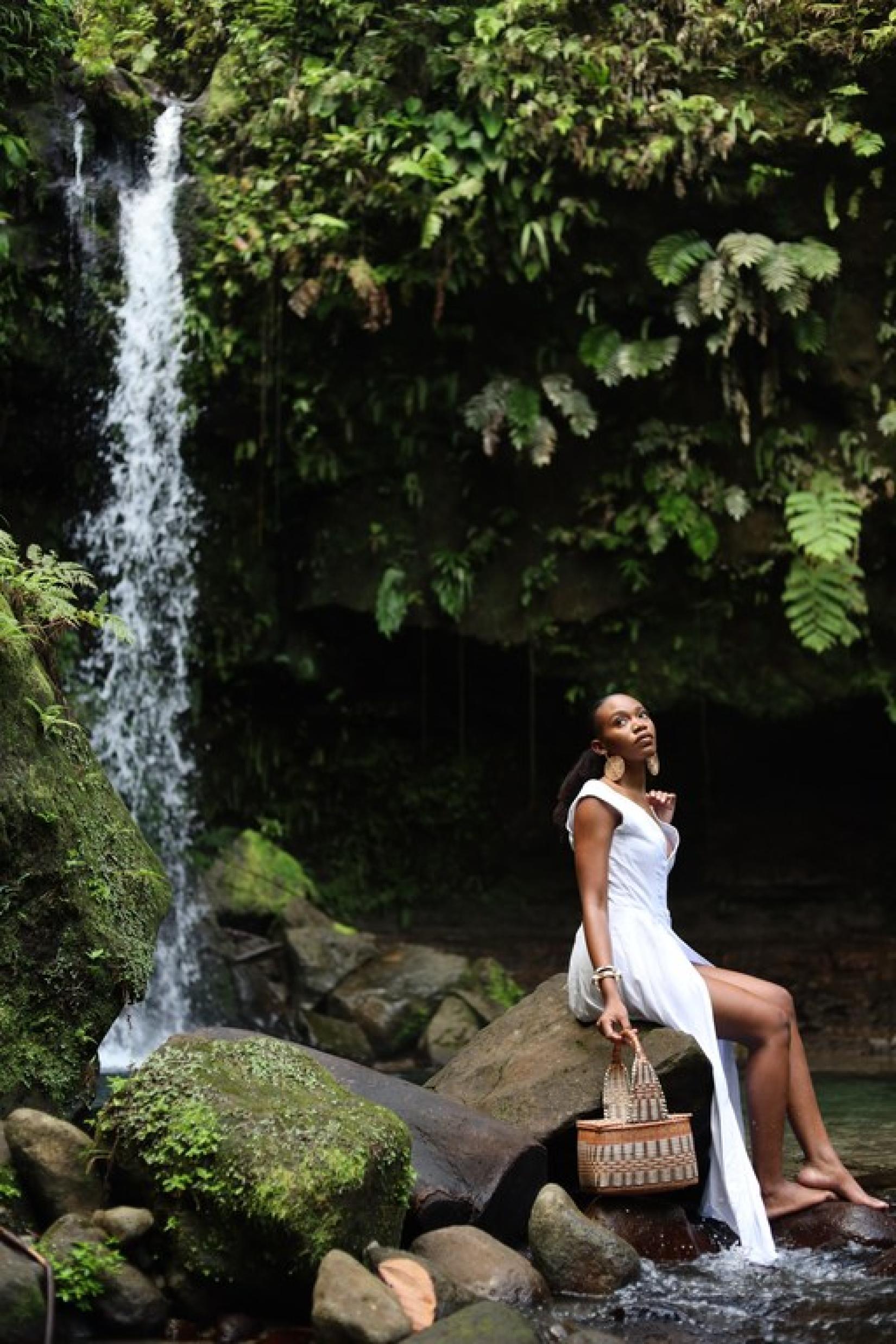 A woman sitting on a rock near a waterfall in a fashionable pose, with a white dress, and styled handbag