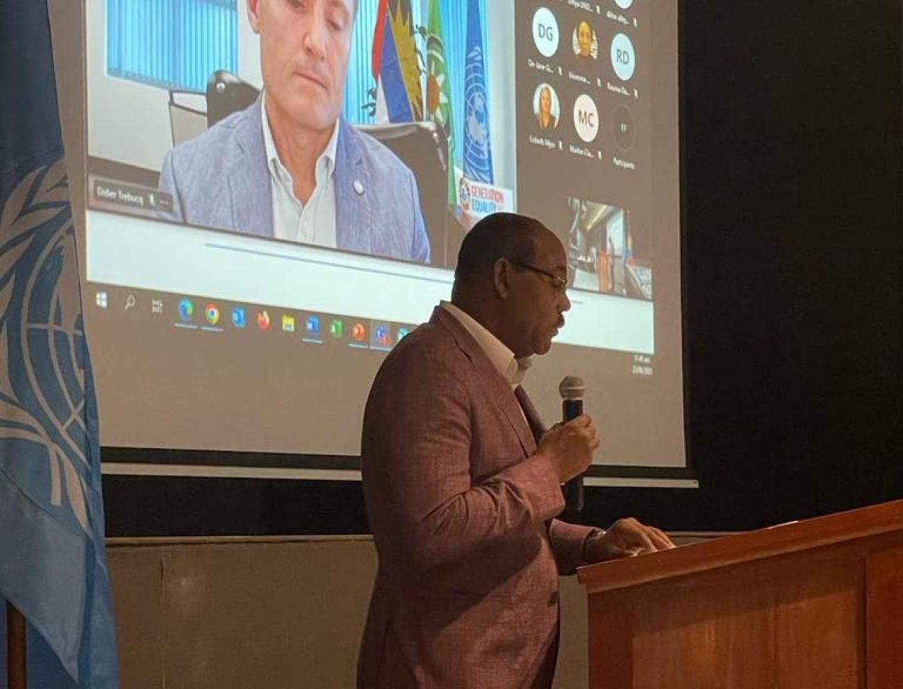 Man standing in blazer at podium with man on screen behind him.