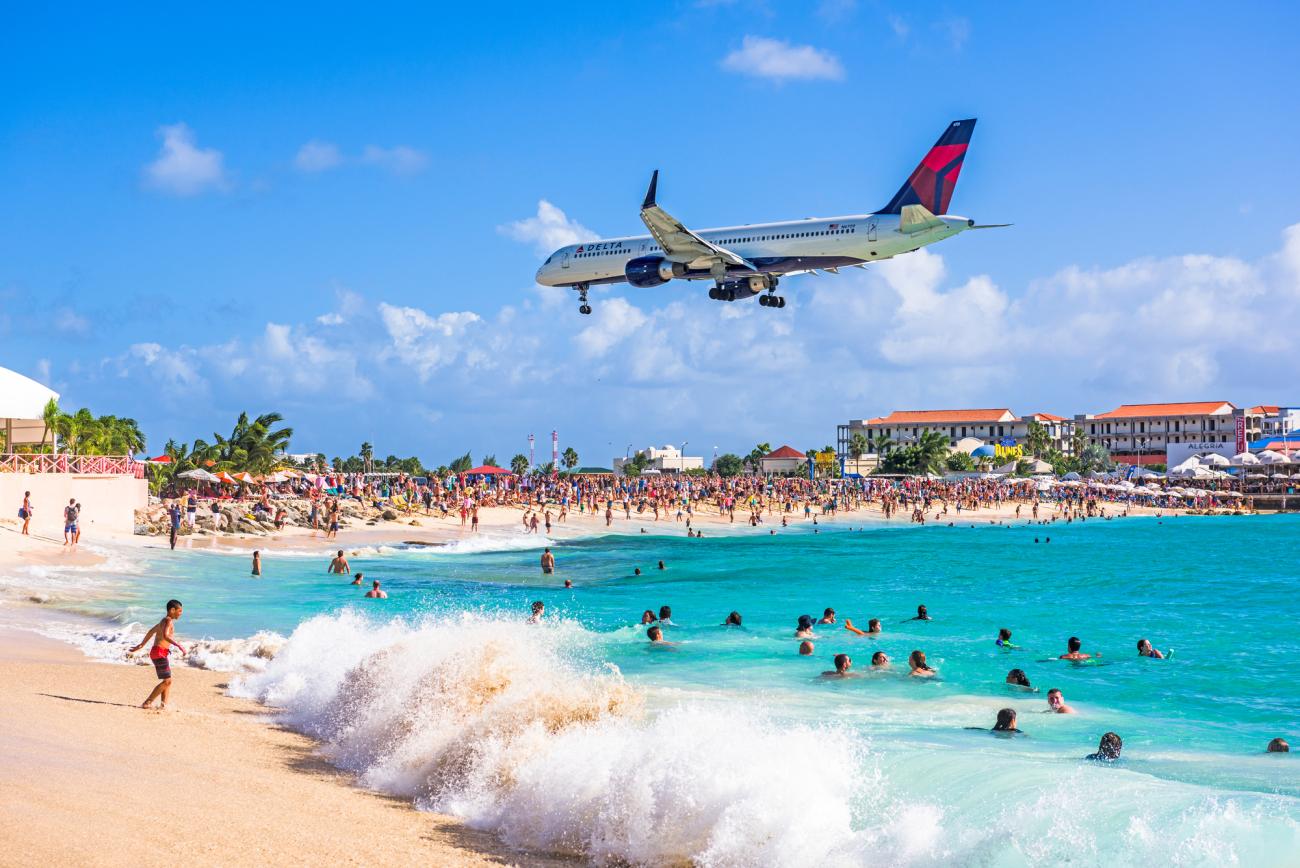 plane flying overhead a beach with persons in the shoreline and in the water 