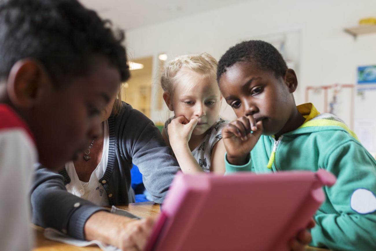 young children in a classroom surround a tablet 