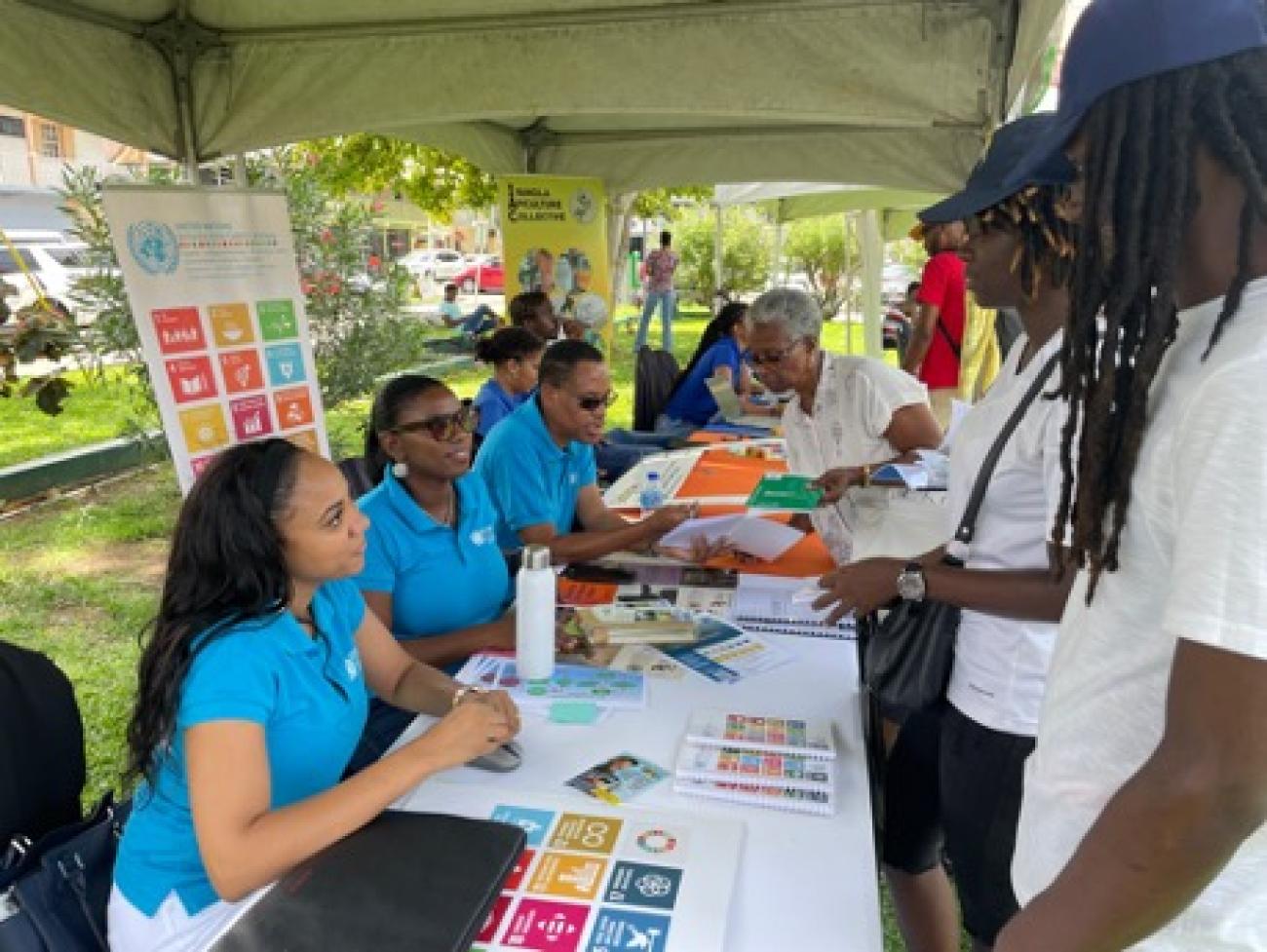 UN Team members sit behind a table backed by an SDG poster interacting with members of the public standing before them.