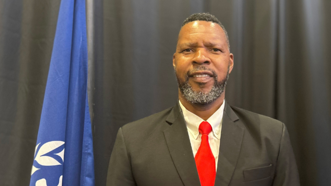 portrait photo of bearded man smiling in suit with red tie and UN Flag in the background