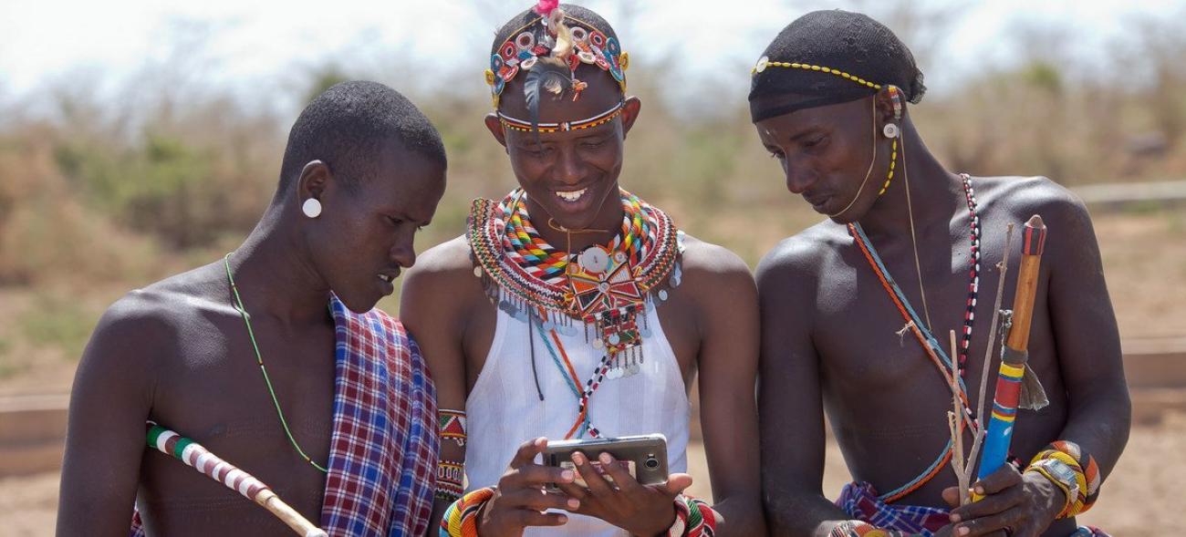 Kenyan pastoralists stand close together in beads and cloths looking into a mobile phone 