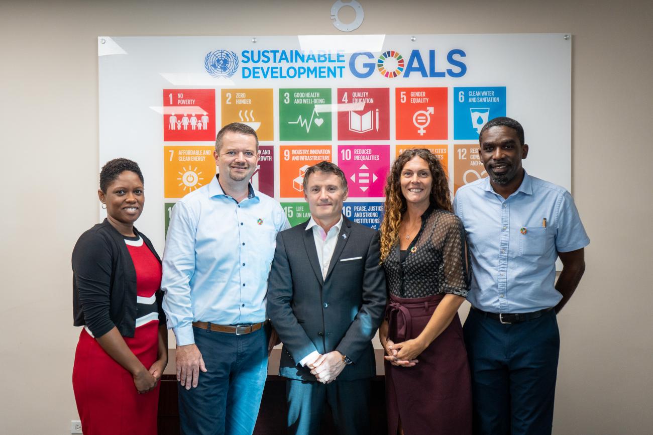 People stand together smiling in a boardroom in front of a Sustainable Development Goals poster