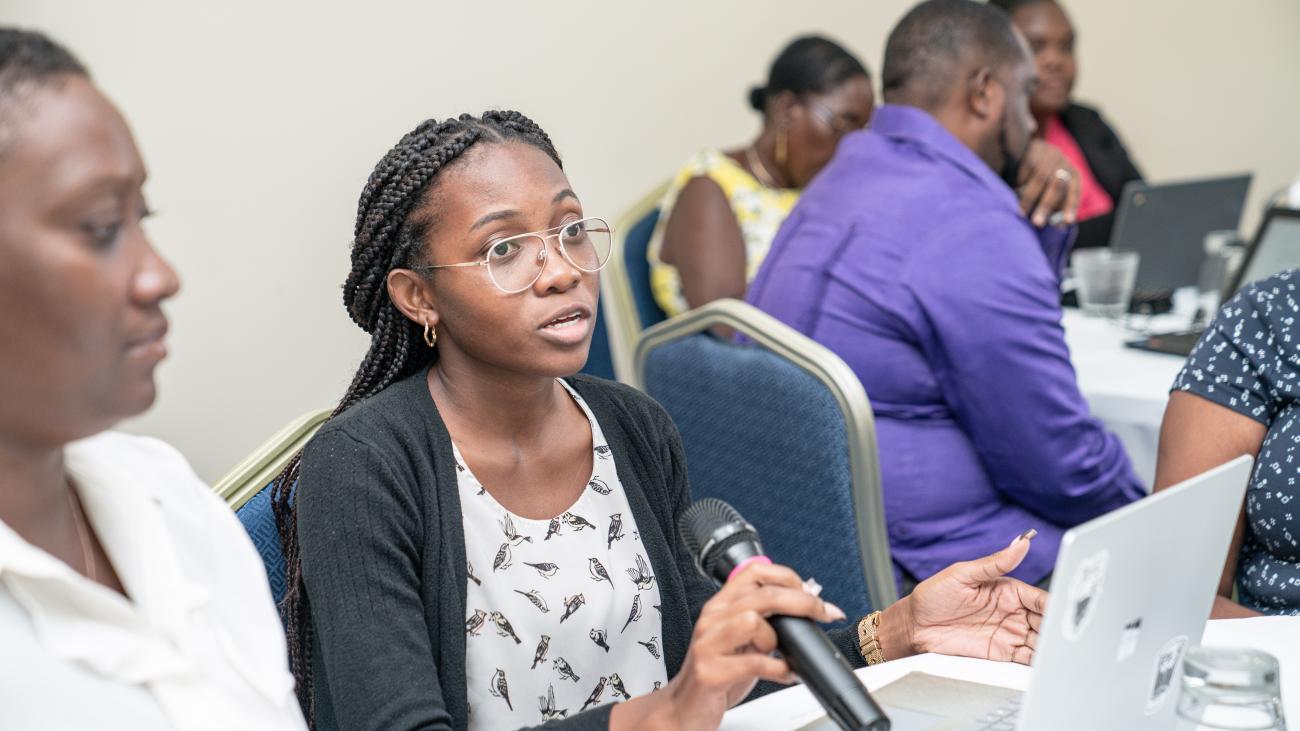 young woman in glasses with braids holds a microphone next to another woman in front of a laptop, speaking in the context of a workshop