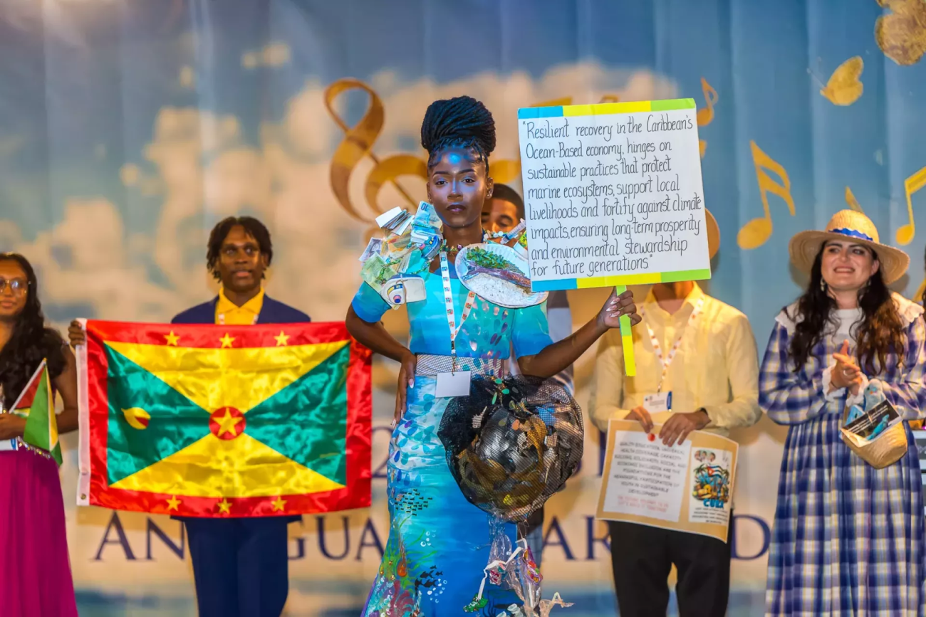 a girl in coastal themed fashion costume holding up a sign on a stage in front of other costumed youth