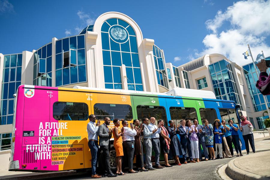 group of 15 persons stand together applauding for a photo against a bus wrapped brightly in the SDG colours, with the UN Seal on a building beyond the bus