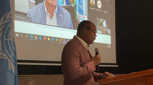 Man standing in blazer at podium with man on screen behind him.