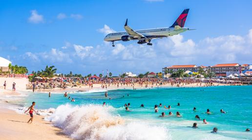 plane flying overhead a beach with persons in the shoreline and in the water 
