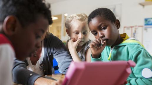 young children in a classroom surround a tablet 