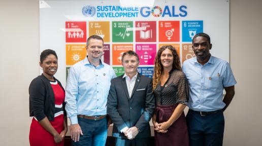 People stand together smiling in a boardroom in front of a Sustainable Development Goals poster