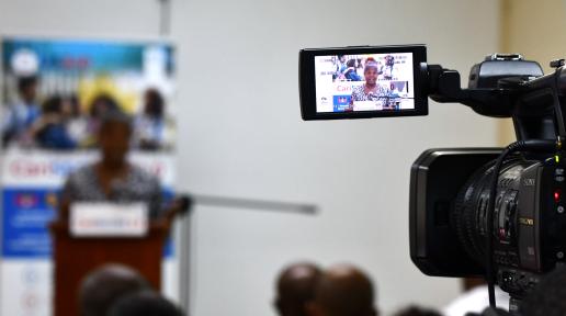 Video camera in focus, capturing a speaker at podium in blurred background