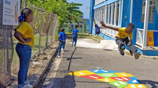 a school boy jumps in midair over a colourfully painted hopscotch board on pavement, while a young school girl looks on