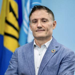 Photo of RC Didier Trebucq standing with arms folded with an SDG Lapel Pin, with the UN, Barbados and OECS flags in the blurred background