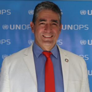 Photo of a man smiling for a photo wearing a red tie, blue shirt, and white jacket with SDG lapel pin