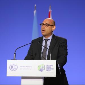 Photo of man in suit and tie and glasses, speaking into an audience